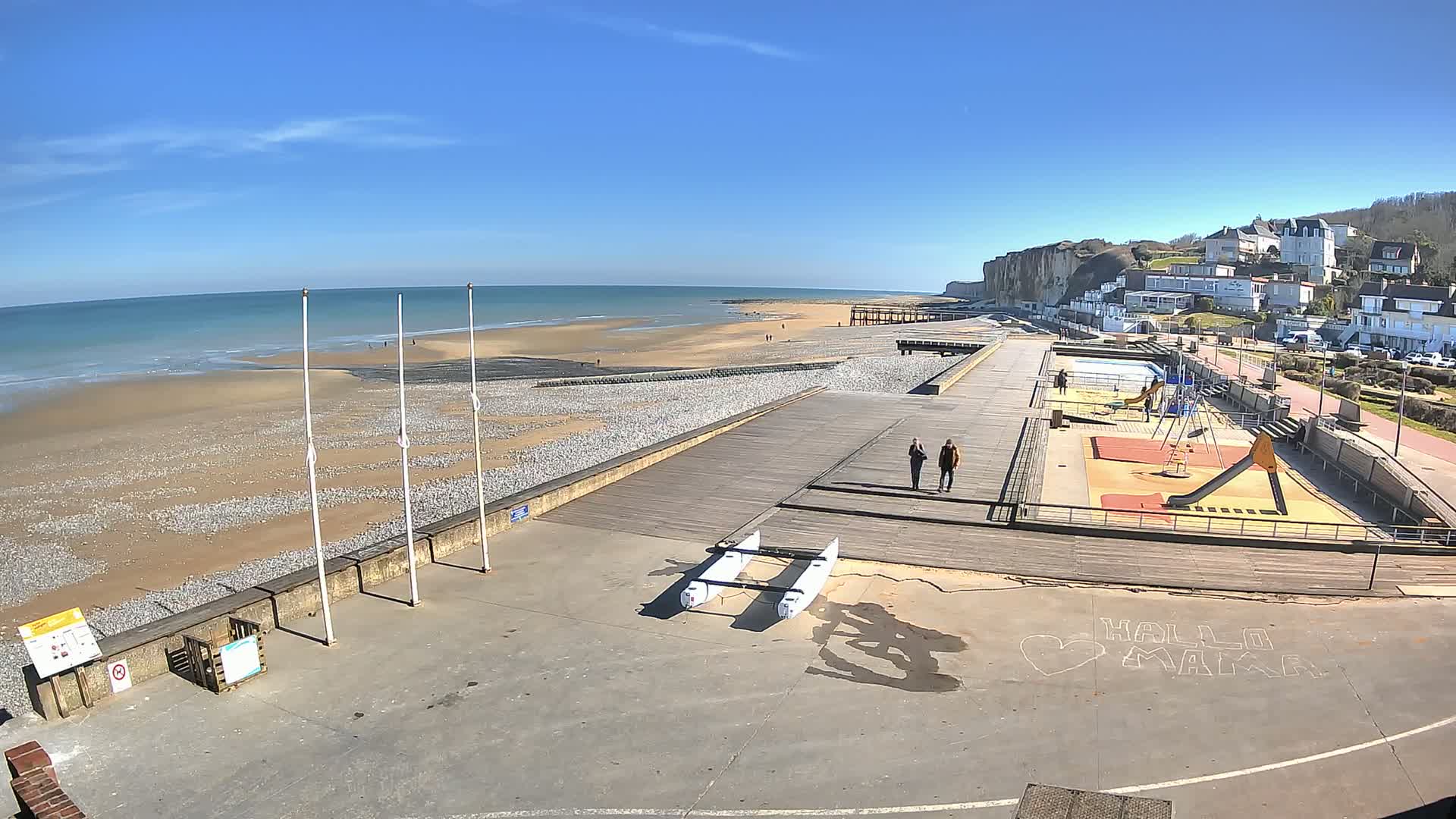A sunny day at a beach with a boardwalk, playground, and a small catamaran, next to white cliffs and a town.