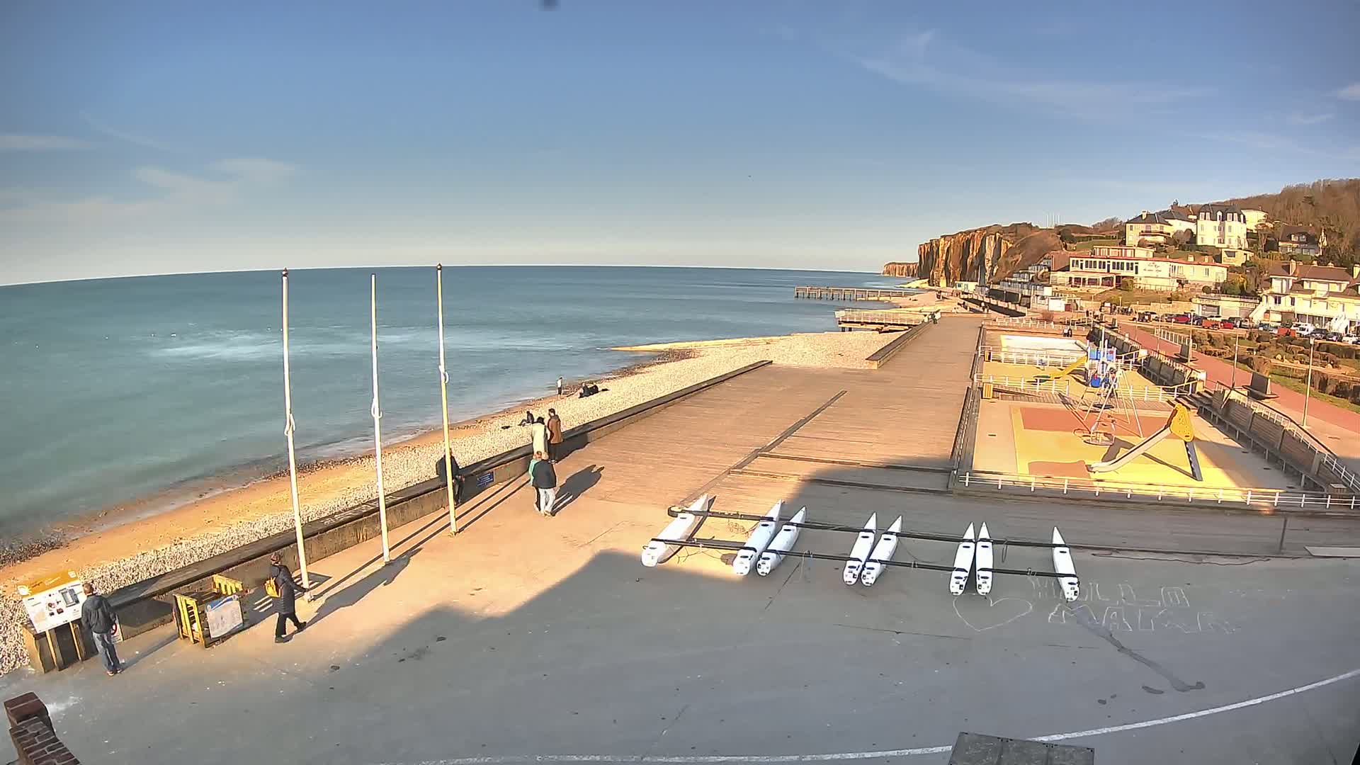 A sunny day at a seaside promenade shows a pebbled beach, calm ocean, a pier, several people walking, a playground, and a line of small boats resting on the paved area.