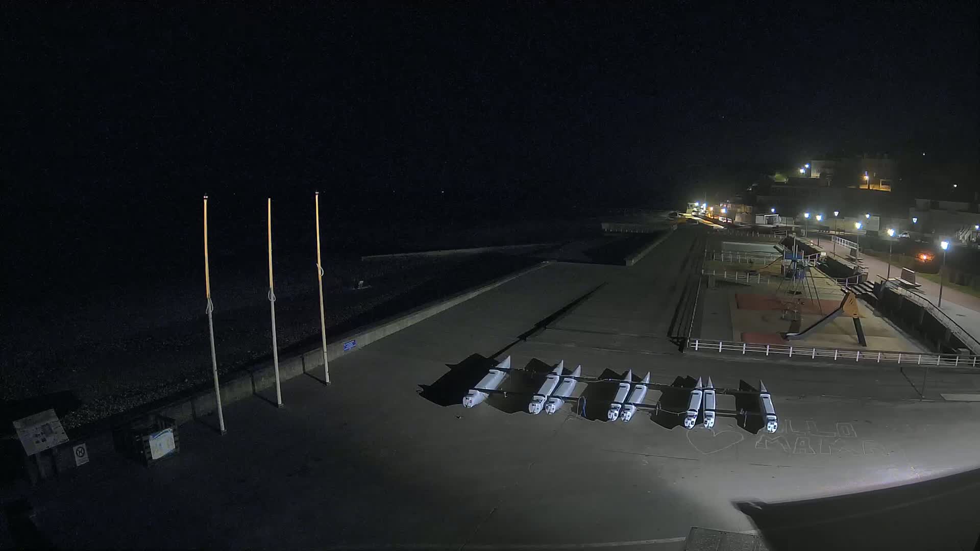 At night, several small boats are lined up on a paved area next to a playground and a dark ocean.