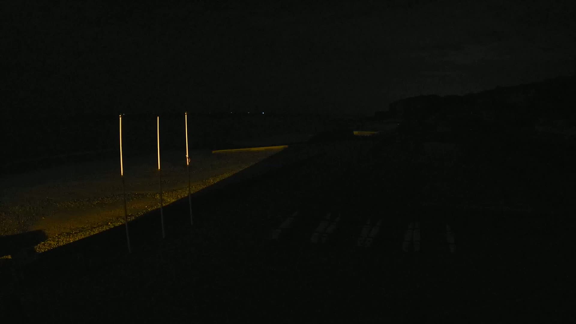Three tall, illuminated poles stand along a dimly lit path at night.