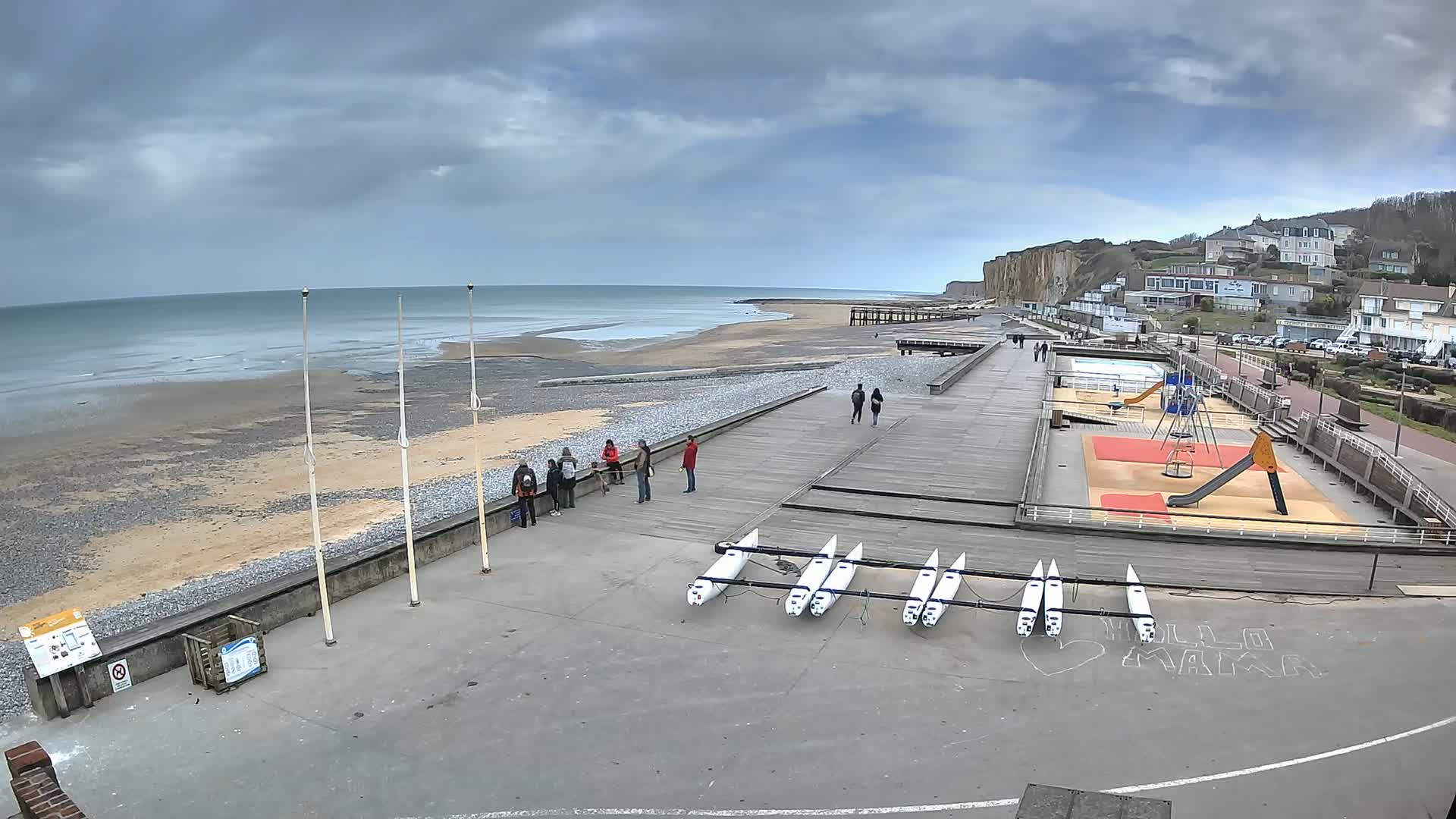 A mostly cloudy day at a beach with a boardwalk, playground, and several small boats lined up on the concrete.