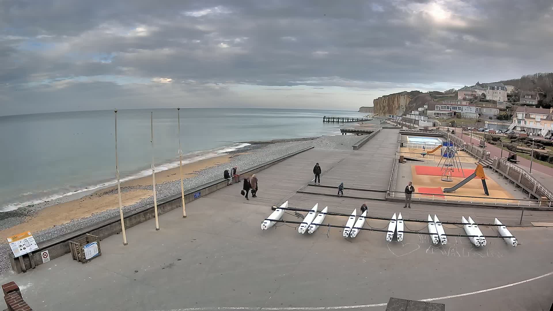 A mostly cloudy day shows a beach with several people, a playground, a pier, and a row of small sailboats lined up on a paved area next to the water.