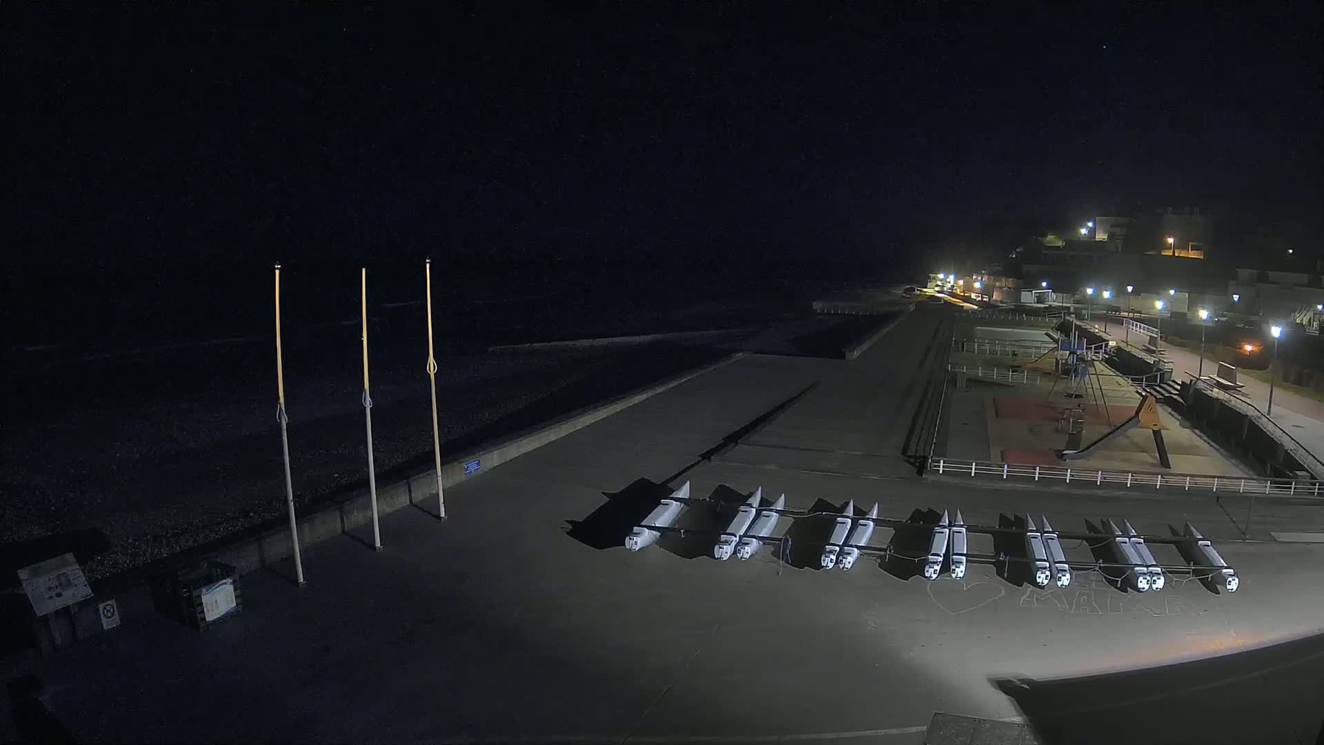 At night, a dark beach with several boats resting on a paved area next to a playground and a walkway with lights is visible under a clear, starry sky.