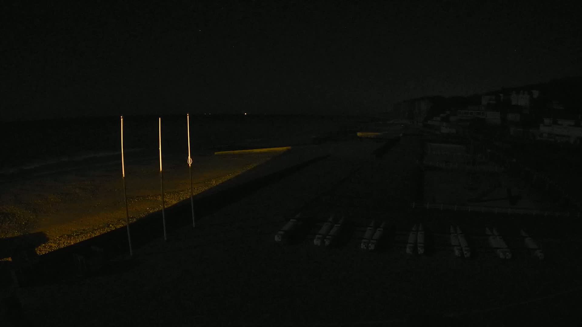 At night, a dark beach with dimly lit tall poles and several long, narrow objects lying on the sand is visible near a dark building.