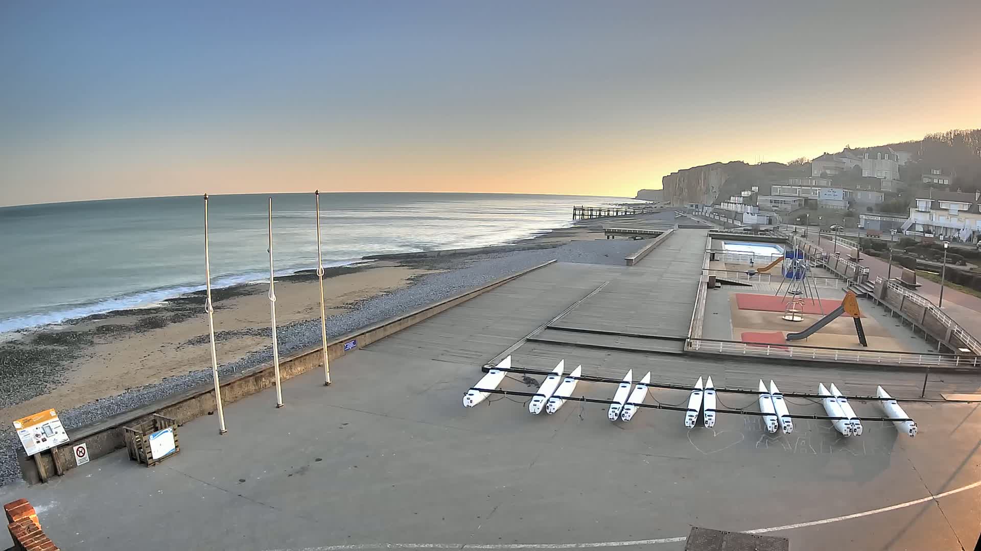 A seaside paved area with several small sailboats rests near a playground, overlooking a calm ocean under a clear, sunrise sky.
