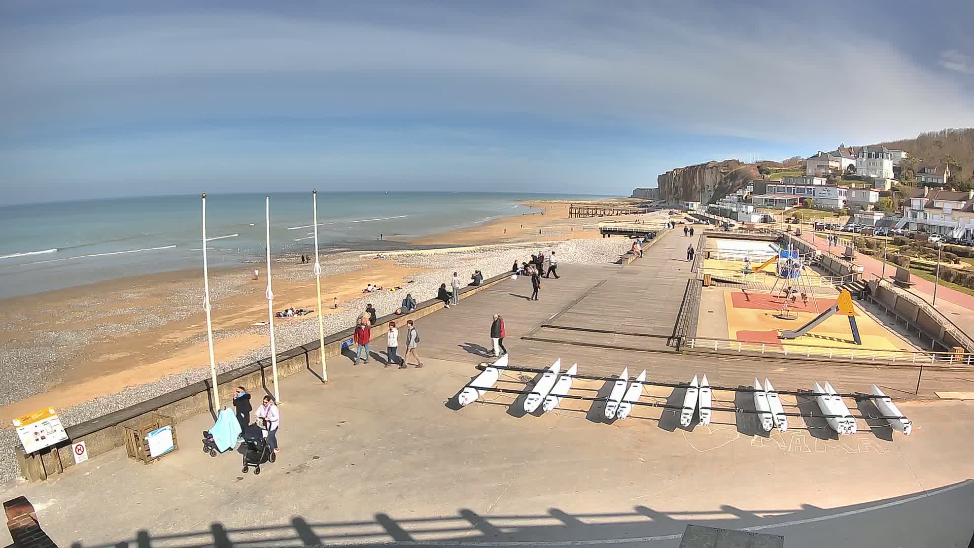 A sunny day at a beach with people relaxing on the sand, walking along a boardwalk with several small boats stored nearby, and a playground visible in the background near white buildings on a cliff overlooking the ocean.