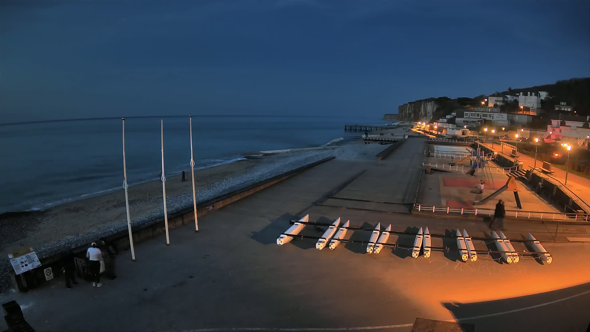 Under a dark blue twilight sky, several small boats are stored on a paved area next to a pebble beach and a seaside town with cliffside buildings.