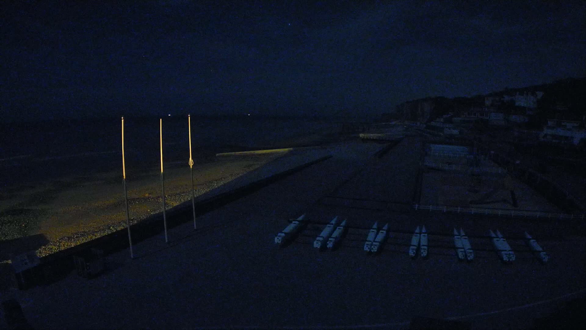 Under a dark night sky, several boats are lined up on a dark beach near a dimly lit town.