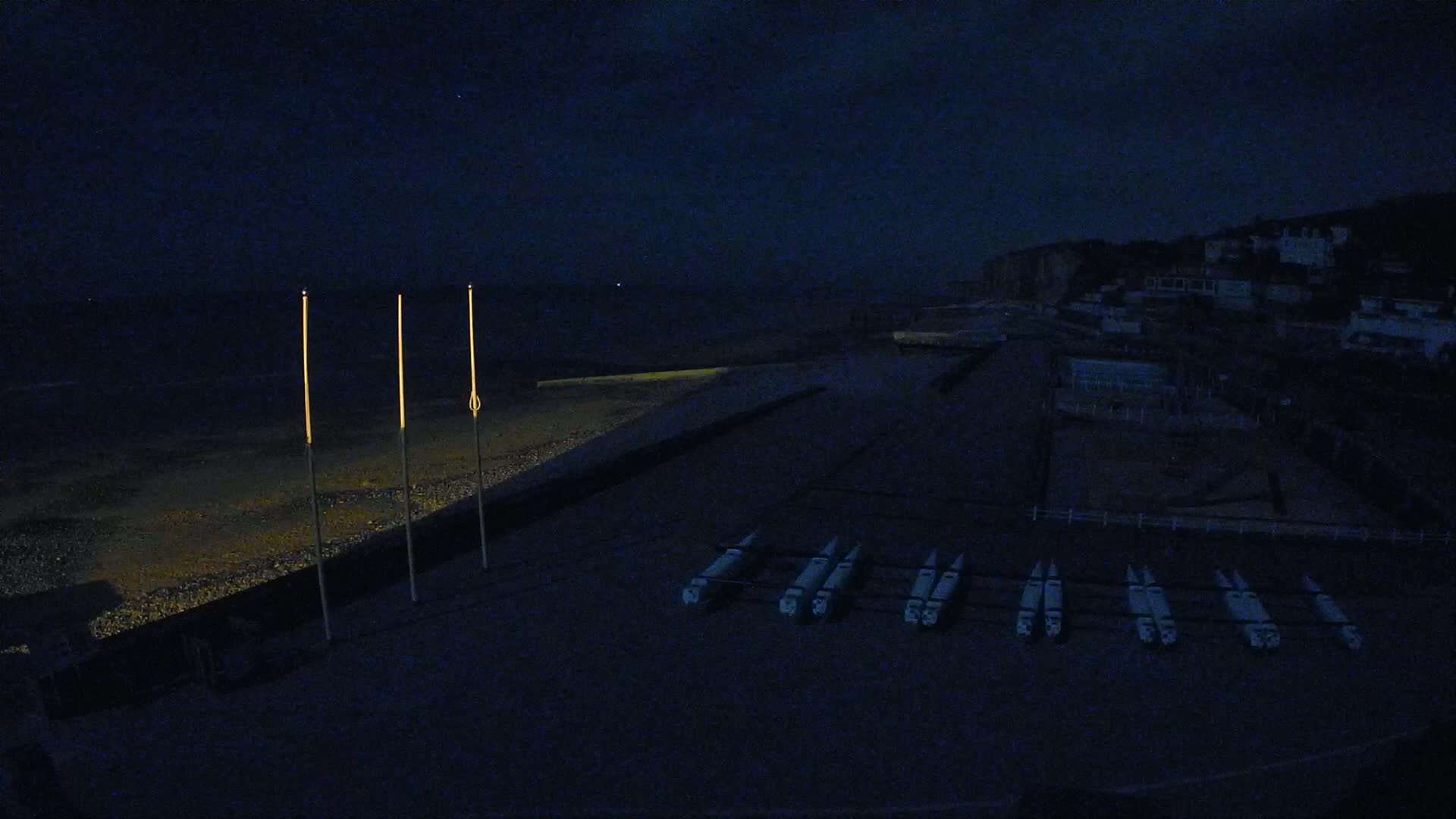 At night, several boats are lined up on a dark beach near a dimly lit town under a dark sky.