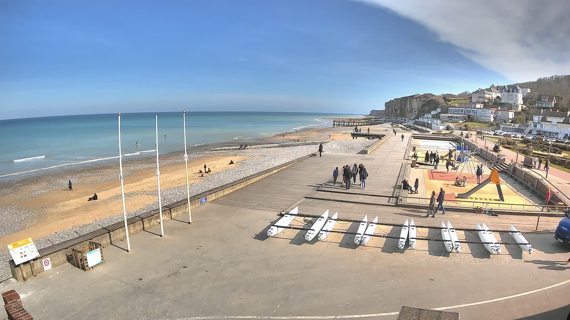 A beach with a wooden boardwalk, a playground, and several small sailboats lined up, under a mostly sunny sky with some clouds.