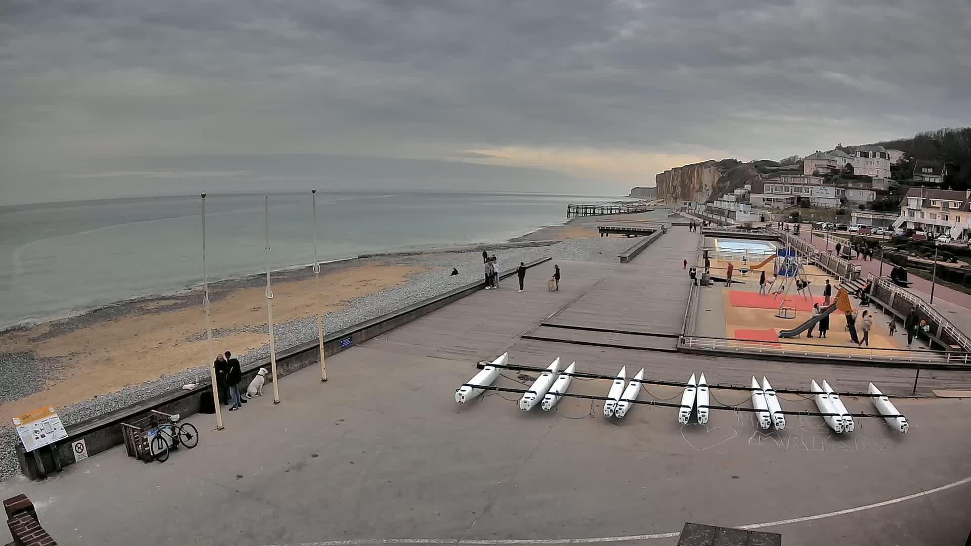 A seaside boardwalk with a playground, several people, a dog, and several boats stored on the ground under an overcast sky.