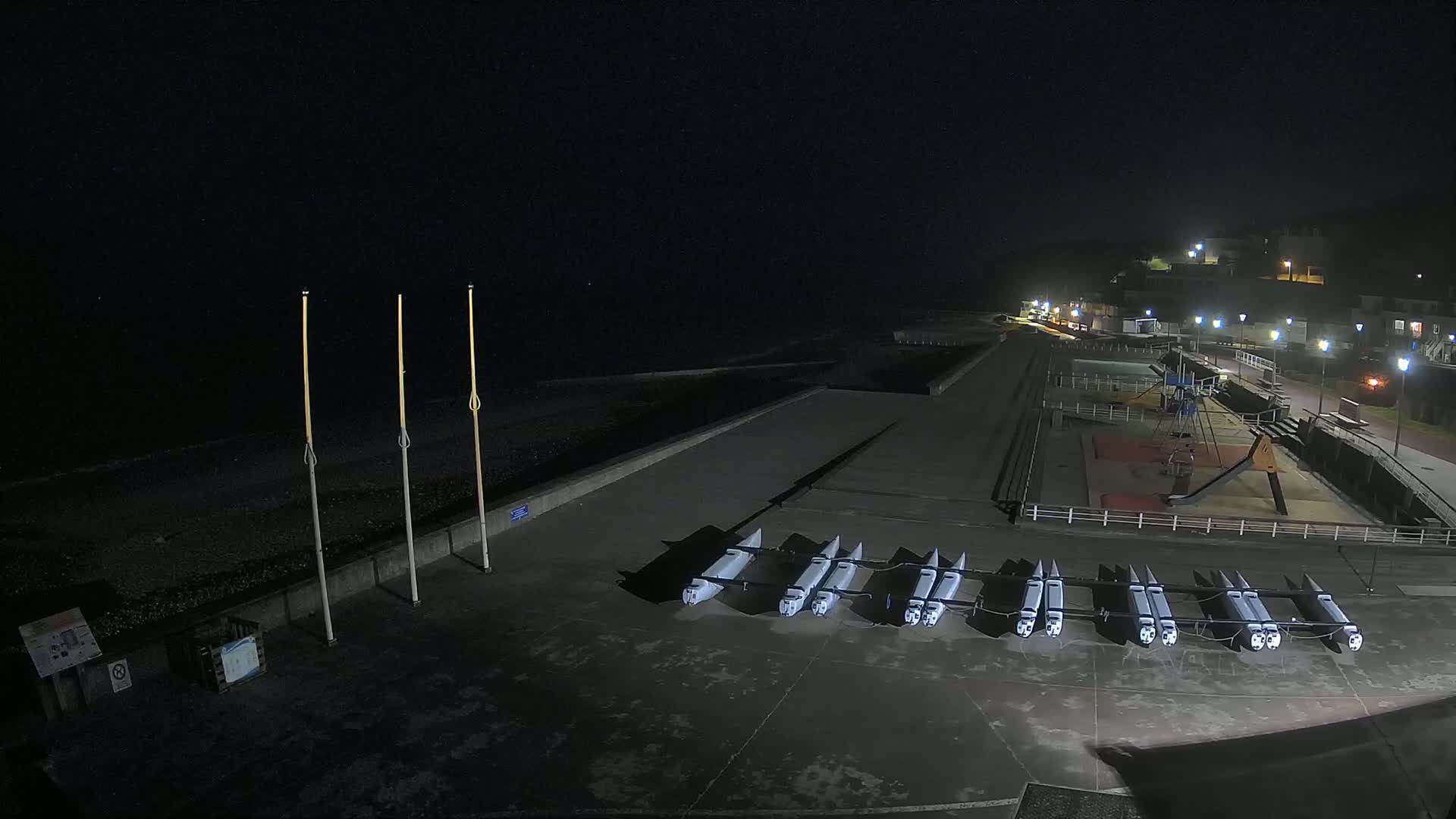 At night, several boats are lined up on a concrete surface near a playground and a pebble beach, under a dark sky.
