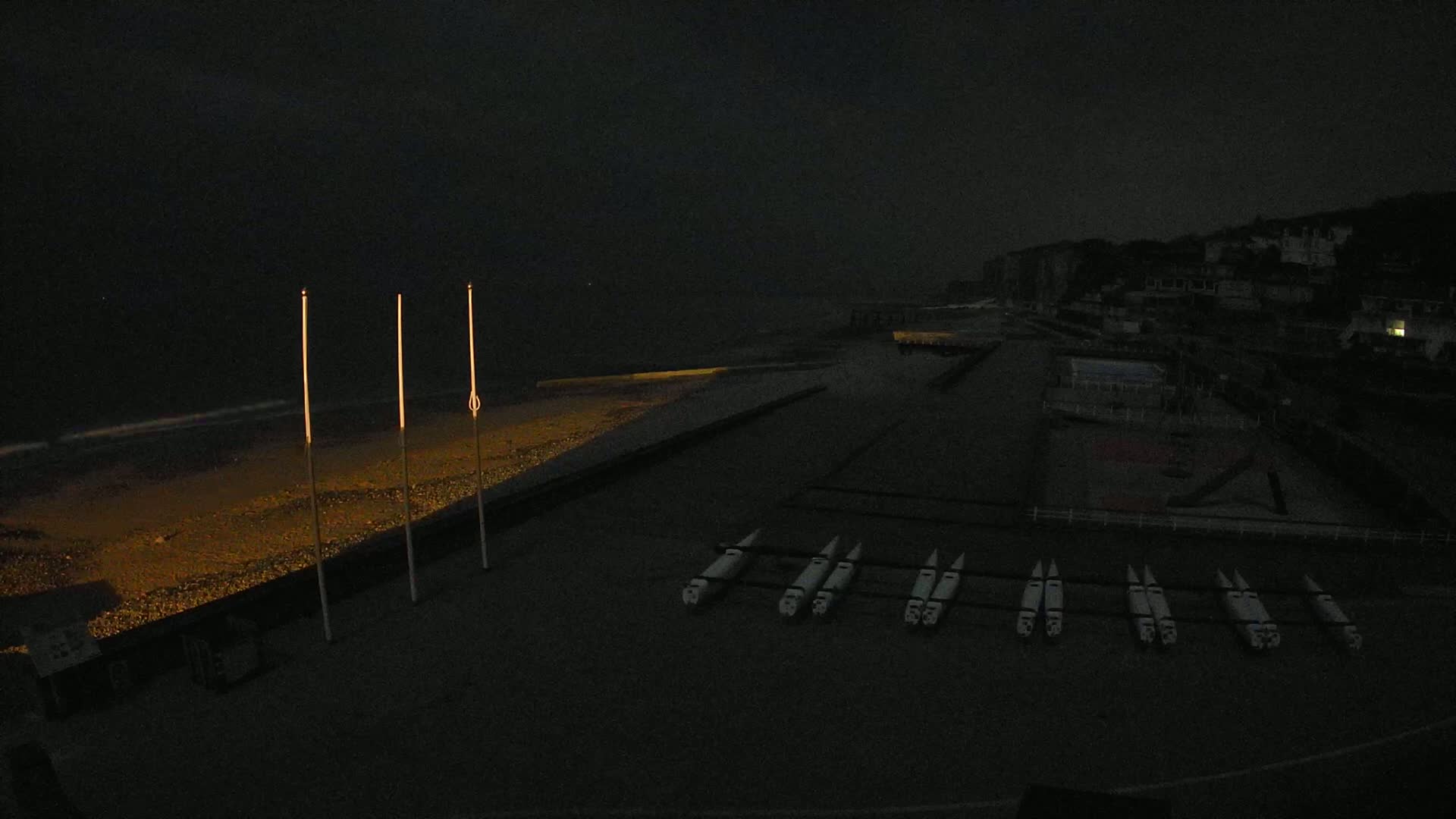 At night, several boats are lined up on a dark beach near a dimly lit town under a dark sky.