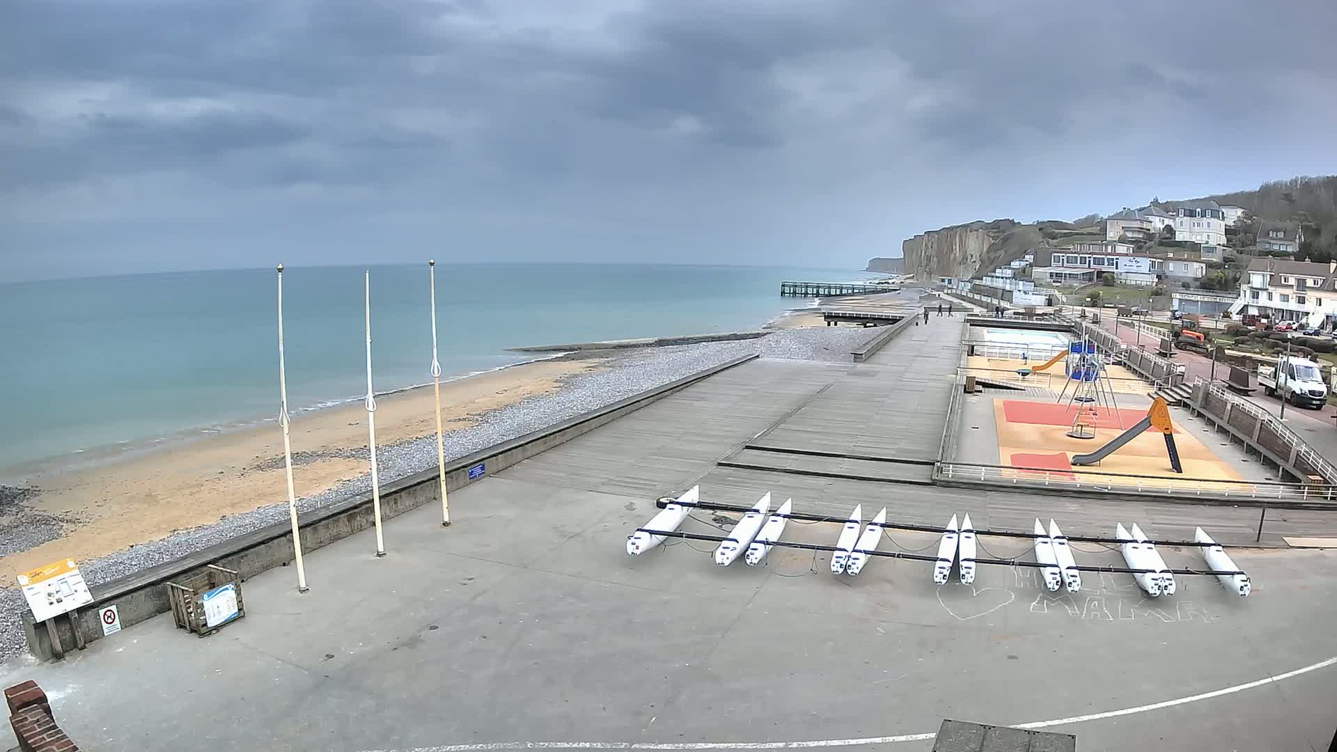 A beach with several small boats and a playground is seen under an overcast sky next to a pier and white cliffs.