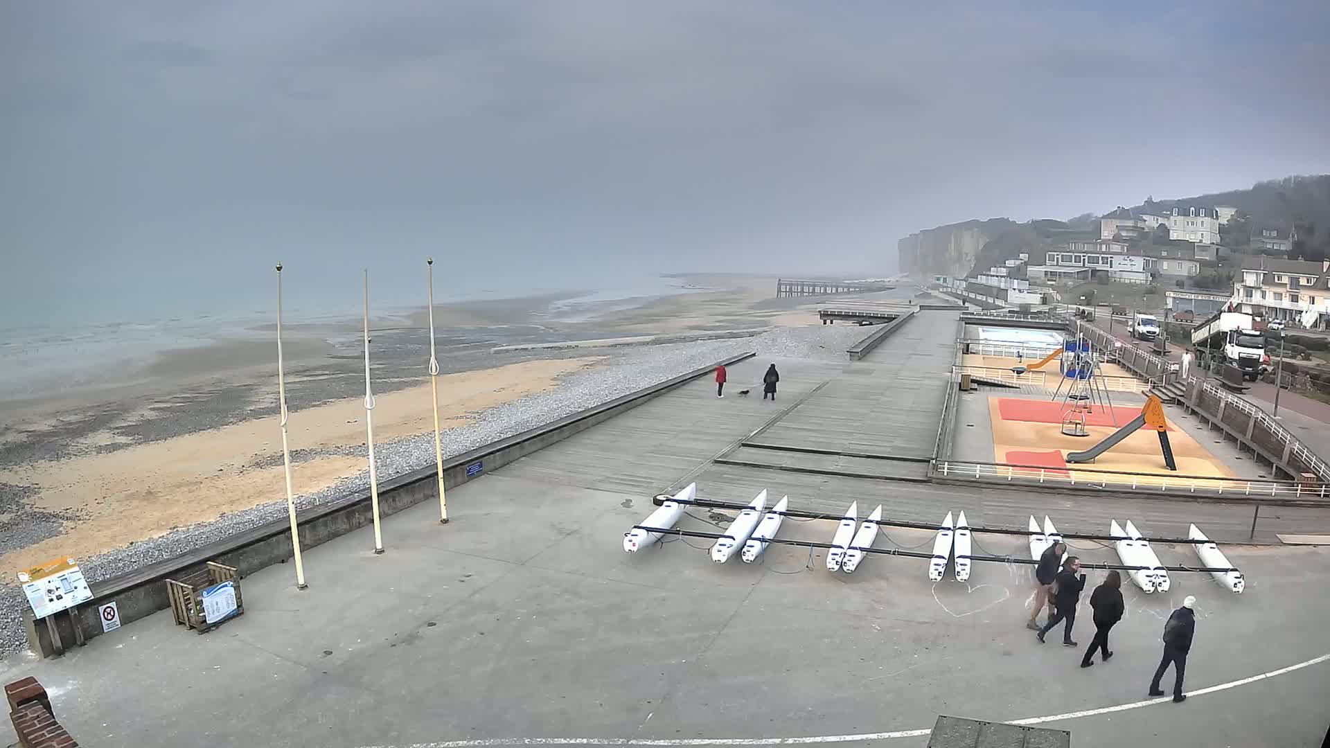 A foggy seaside boardwalk features several small sailboats, a playground, and a few pedestrians, with white cliffs visible in the distance.