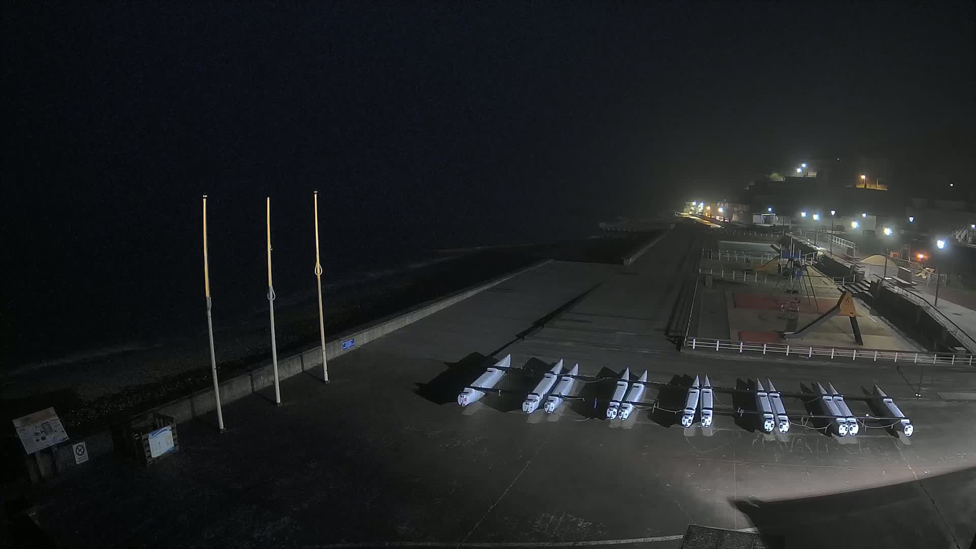 At night, under a dark, possibly foggy sky, several white boats are lined up on a concrete surface near a playground and a seawall.
