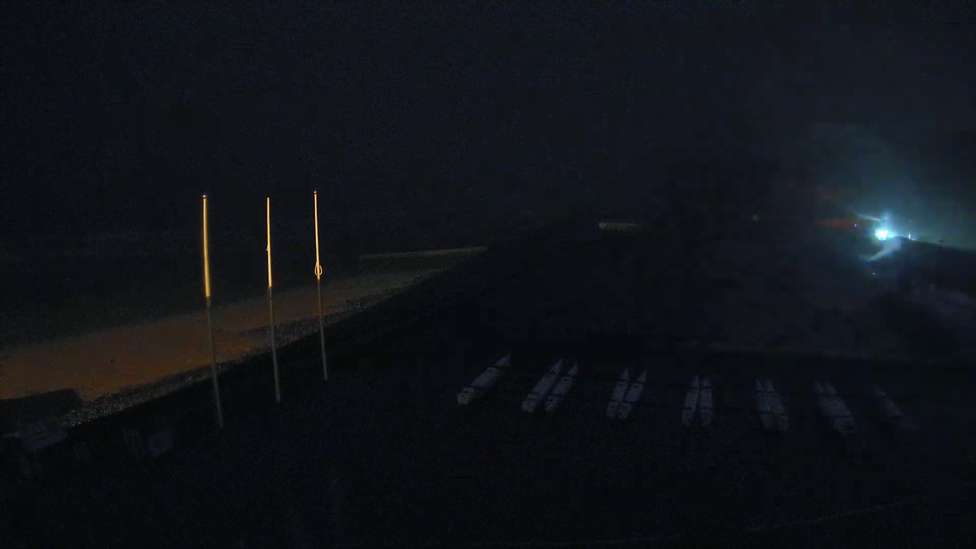 Several boats are docked at a dark shoreline at night in foggy conditions, illuminated by a few tall lights.