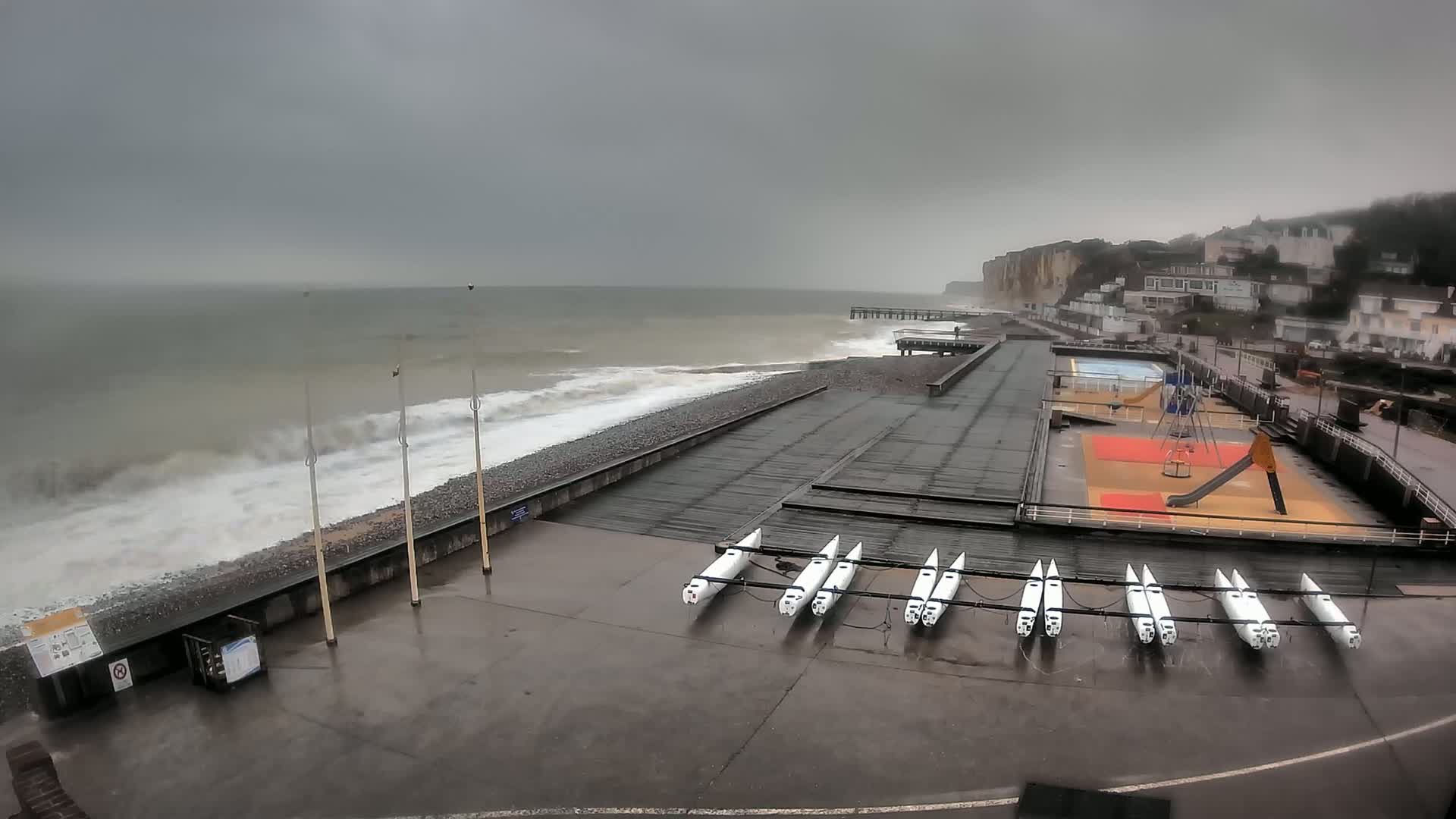 A stormy grey day at a seaside town shows a rocky beach with crashing waves, a pier, a playground, and several small boats stored on the shore.