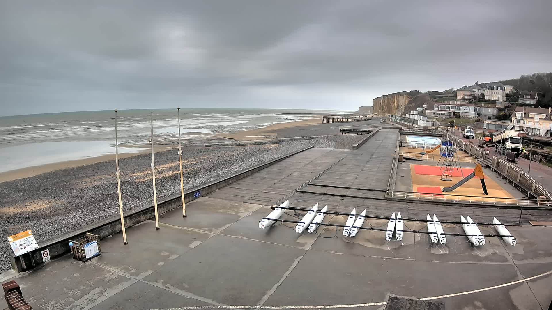 A beach with a playground and several small boats stored on the shore under an overcast sky with a choppy sea.