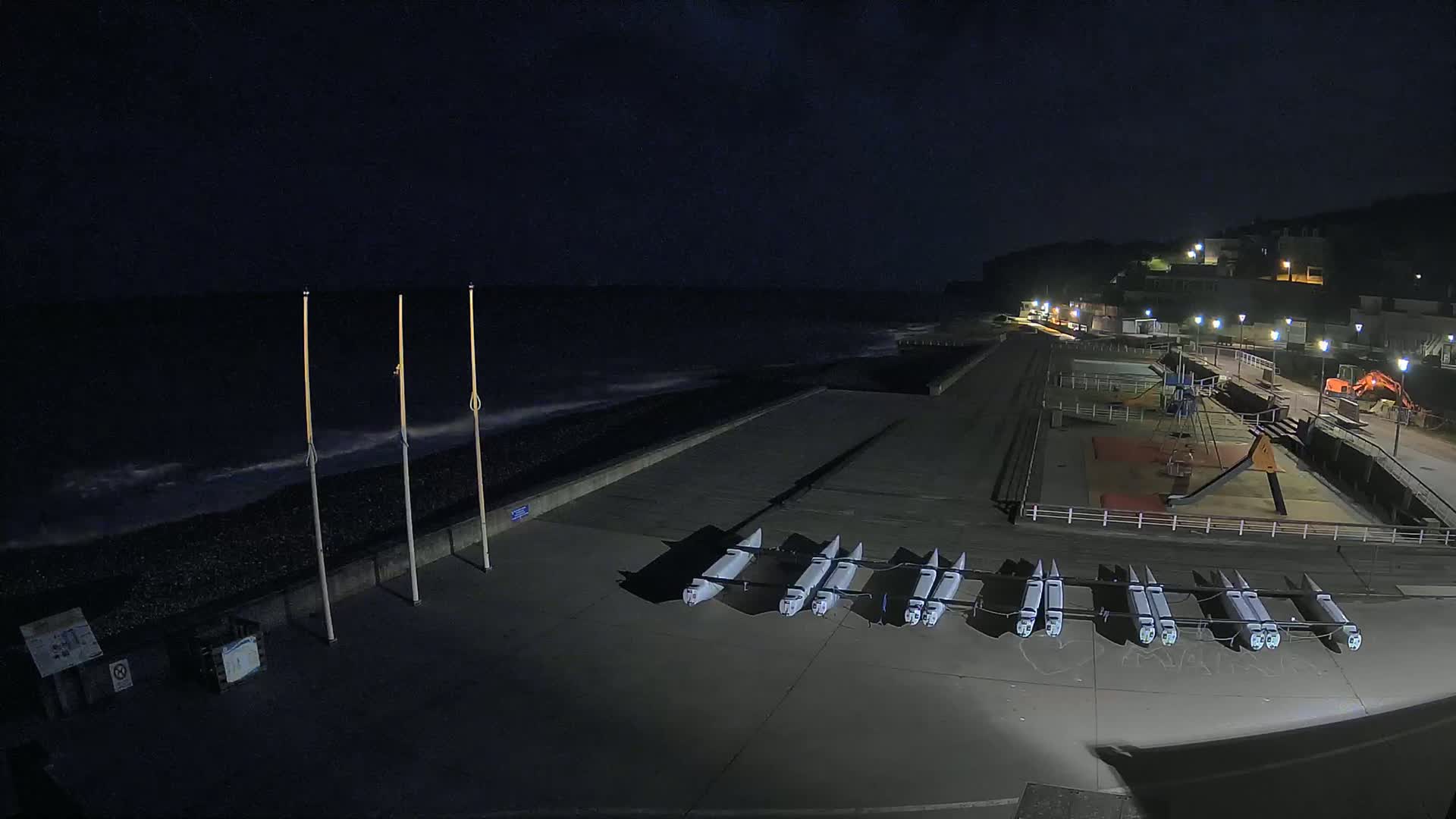 At night, several small boats are lined up on a paved area next to a dark, wavy ocean, with a playground and lights visible in the background.