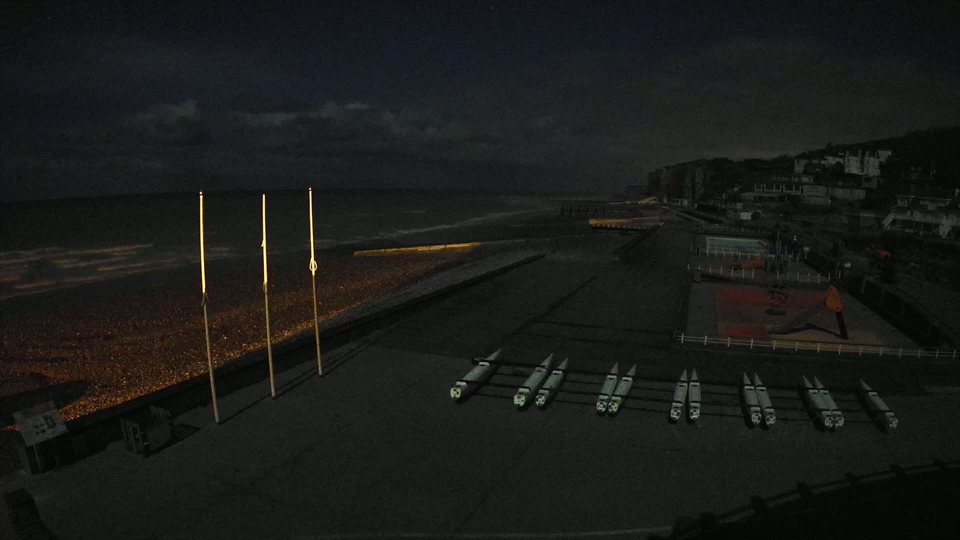 At night, under a mostly cloudy sky, several rows of boats are lined up on a dark paved area next to a pebble beach and a calm sea.