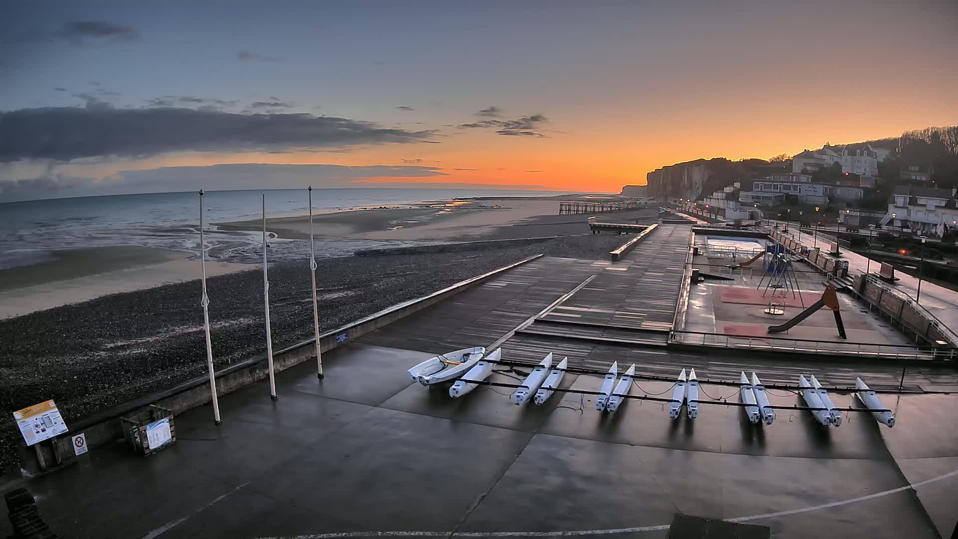 A wet, paved waterfront area with several small sailboats, a playground, and a swimming pool overlooks a rocky beach and ocean at sunset under a partly cloudy sky.
