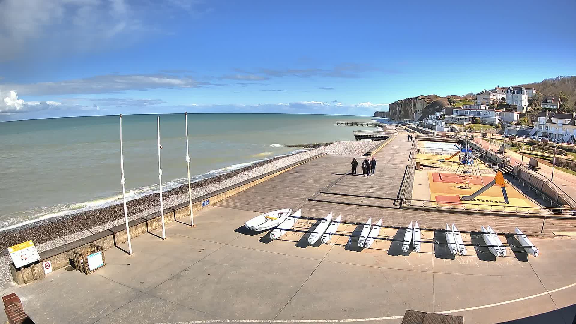 A boardwalk overlooks a pebble beach and calm ocean under a partly cloudy sky, with several small sailboats stored nearby, a playground, and a small group of people walking in the distance.