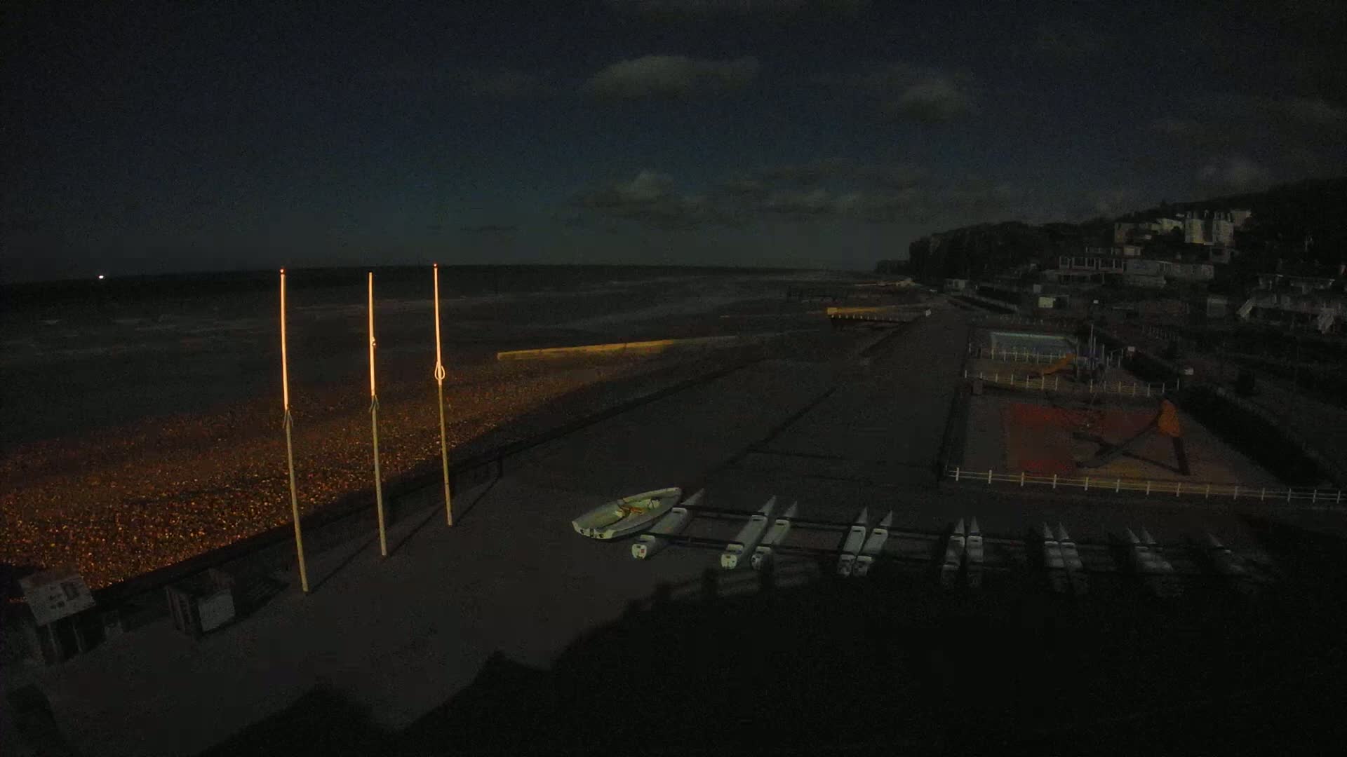 A dark, nighttime beach scene shows several small boats and tall poles on a pebbly shore under a mostly cloudy sky with a slightly wavy sea.
