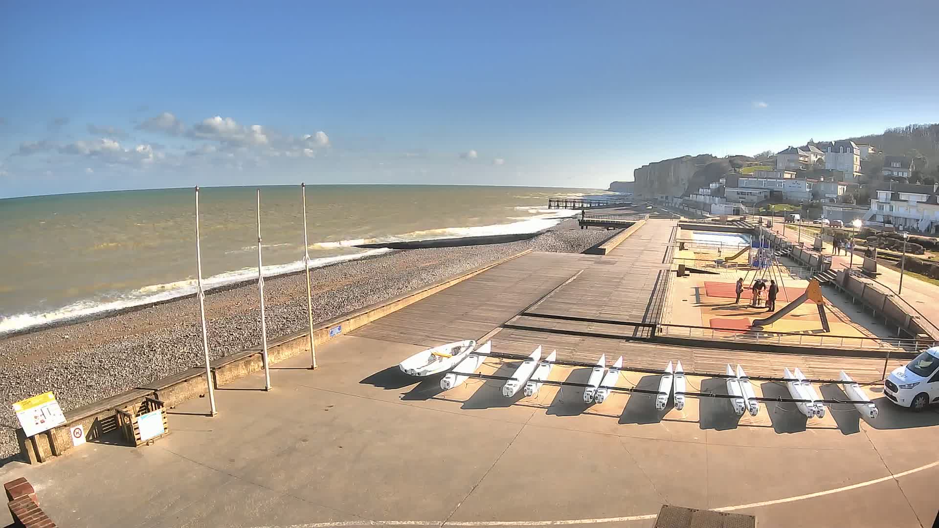 A pebble beach with a wooden boardwalk, small boats, a playground, and buildings overlooking a wavy ocean under a mostly sunny sky.