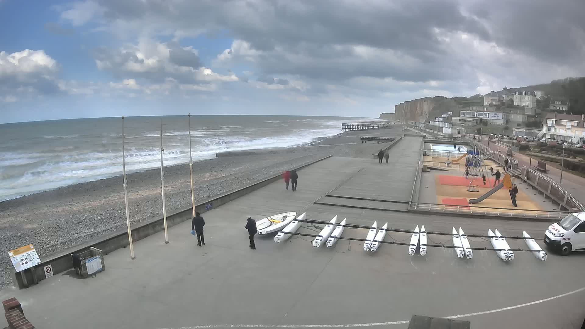 A rocky beach with choppy waves under a cloudy sky is next to a paved area with several small sailboats, a playground, and a few people walking.