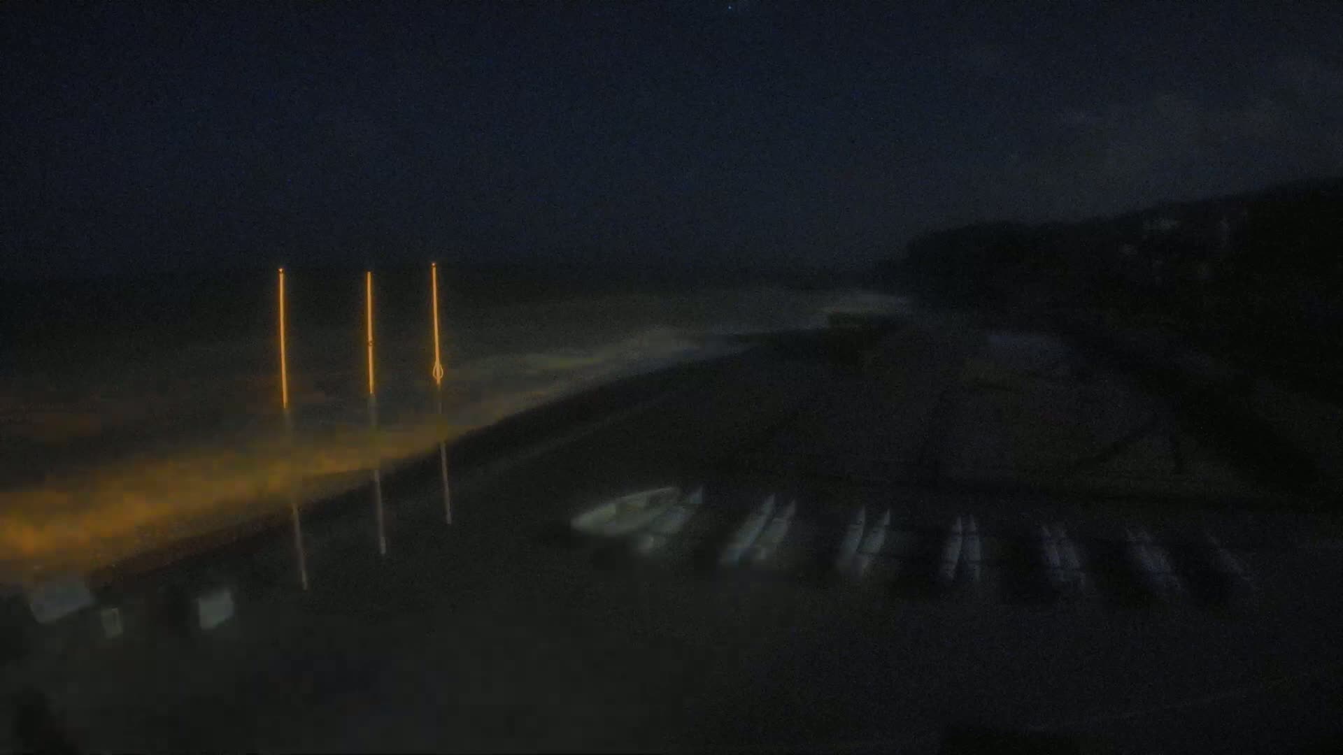 Three tall, illuminated poles stand near a dark, nighttime shoreline where several small boats are moored, with a faintly visible ocean and a dark sky above.