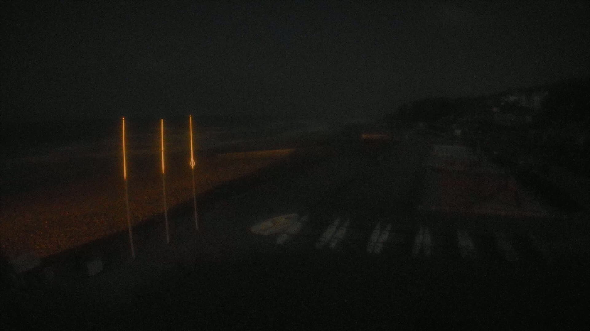 Three tall, thin, orange lights stand on a dark beach at night, with several dark objects visible in the water nearby.