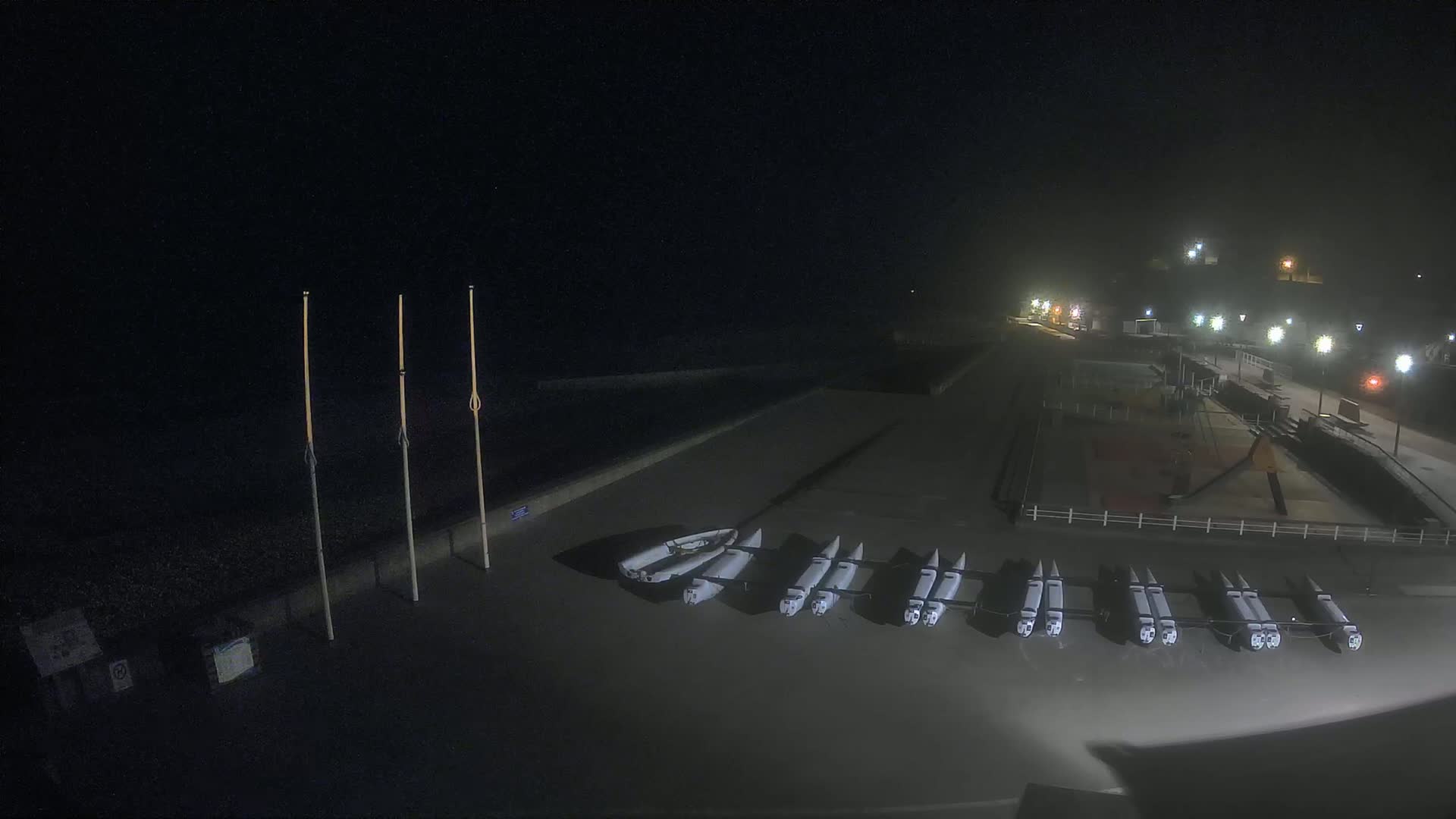 At night, under hazy conditions, several small white boats are stored on a dark paved area next to a dimly lit waterfront.