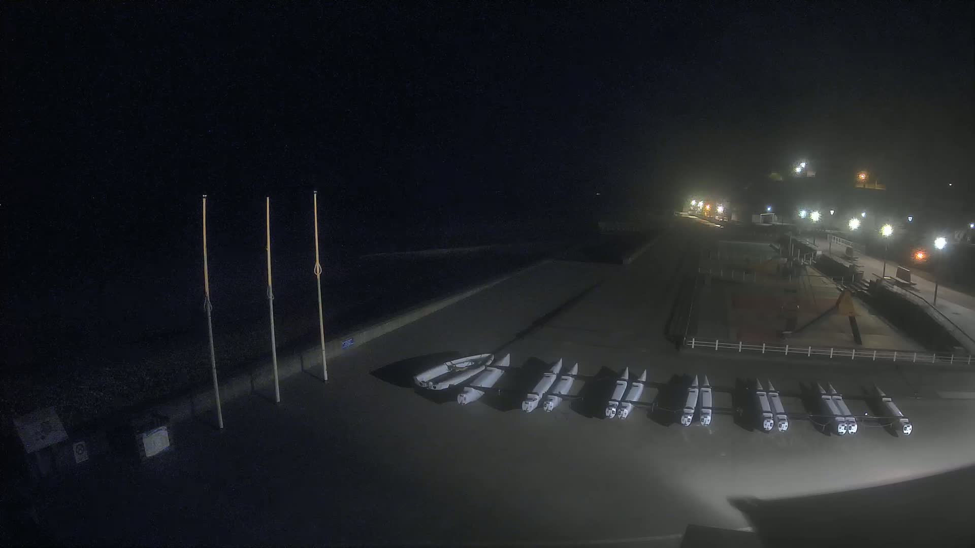 At night, several small boats are stored on a dark, wet surface near a paved area with lights and tall poles.