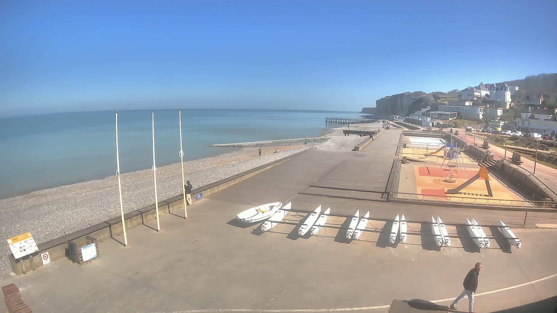 A pebble beach and boardwalk overlook a calm sea under a clear blue sky, with several small boats stored nearby and a playground visible in the upper right.