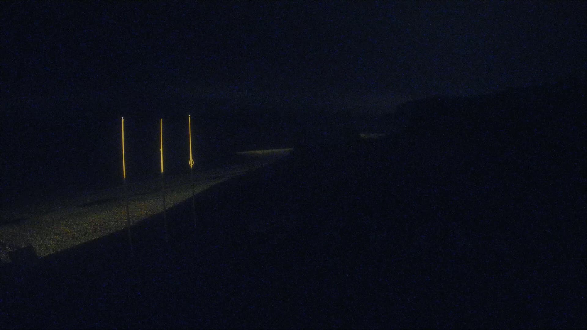 Three tall, slender lights illuminate a dark, possibly paved, surface at night.