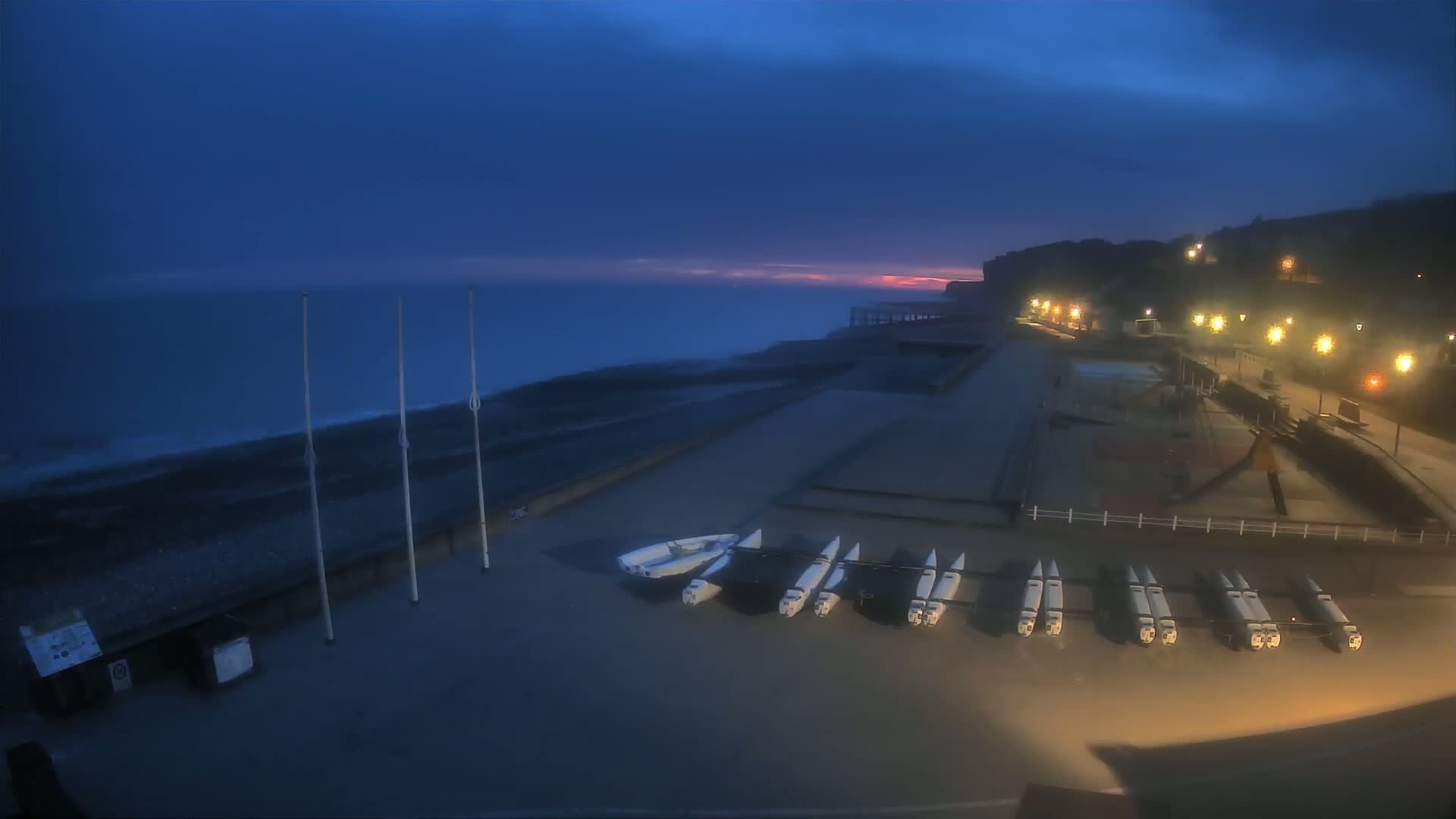 A dark, twilight scene shows a beach with several small sailboats parked on the shore, under a mostly cloudy sky with a hint of pink at the horizon.