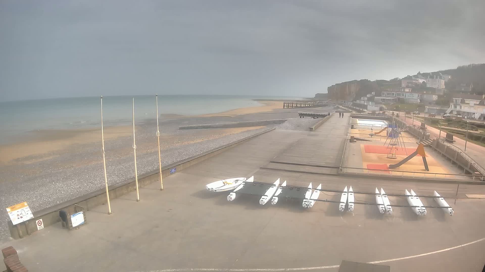 A seaside area with overcast skies shows a beach, a boardwalk, several small sailboats, and a playground next to residential buildings on a cliff.