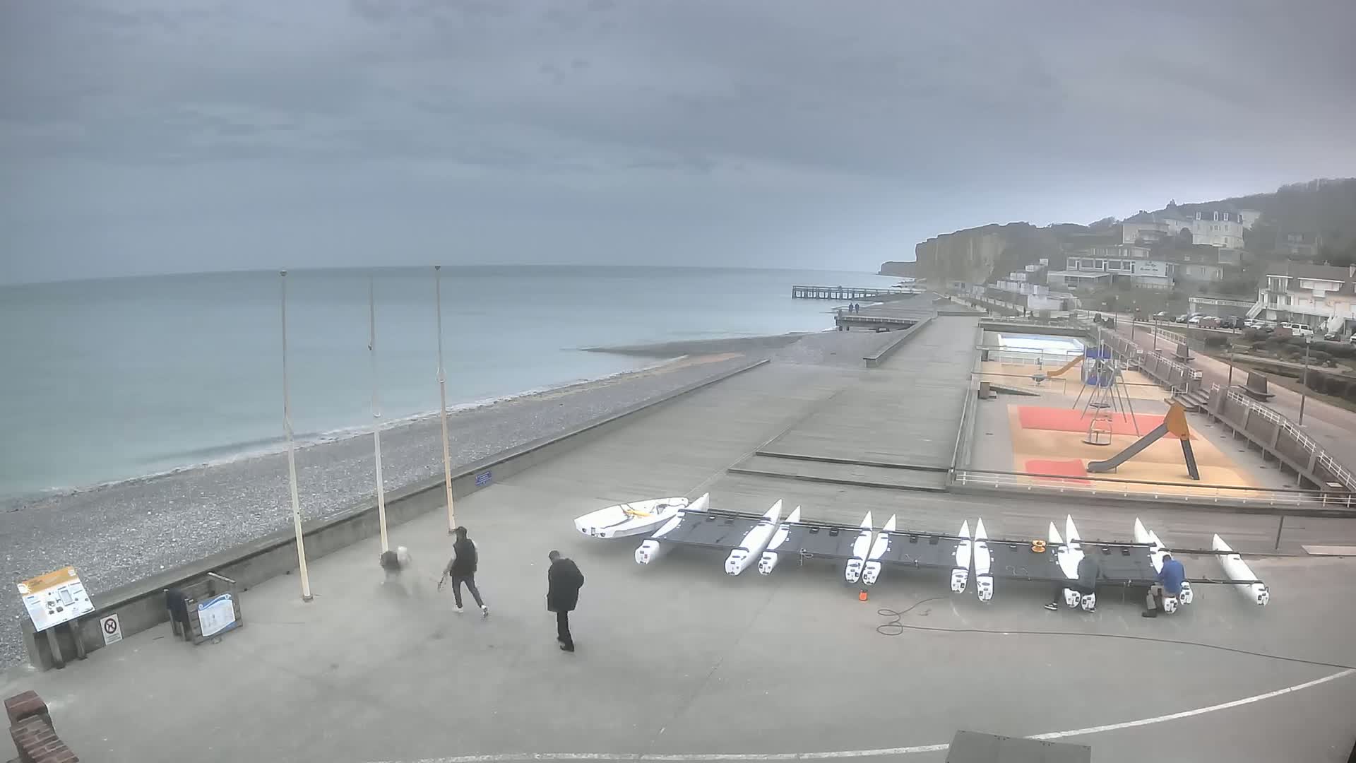A gray, overcast day at a seaside promenade shows several people walking near a beach with a row of small sailboats, a playground, and buildings in the background.