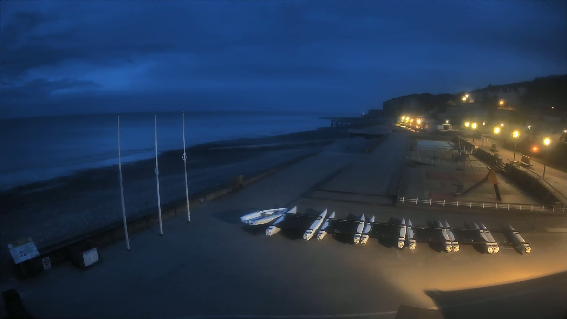 Under a dark blue, twilight sky, several small sailboats are lined up on a paved area next to a pebble beach and a dimly lit seaside promenade.