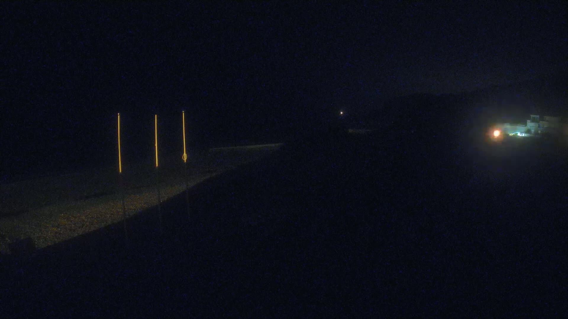 At night, three illuminated poles stand on a dark, pebble-covered beach near a dimly lit building.
