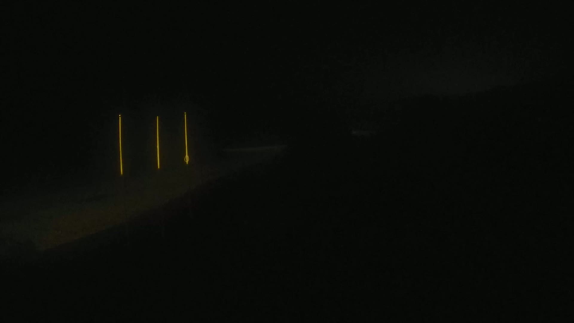 Three tall, thin, yellow lights stand against a dark, nighttime landscape.