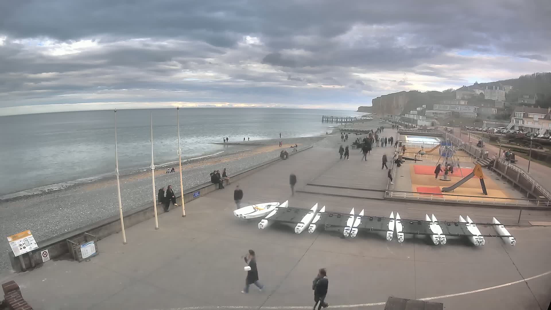 A mostly cloudy day overlooks a pebble beach with people walking along a promenade, small boats stored on the ground, and a playground visible near buildings and a pier in the distance.