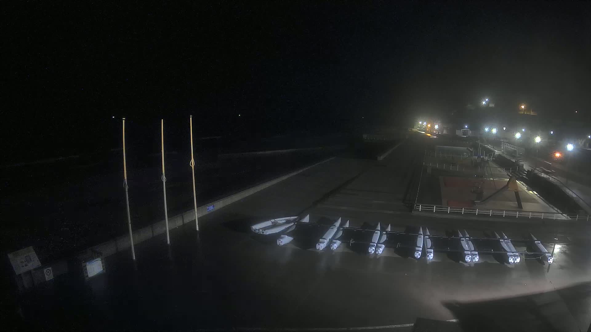 At night, under foggy conditions, several small boats are lined up on a dark, wet waterfront near a lighted promenade.