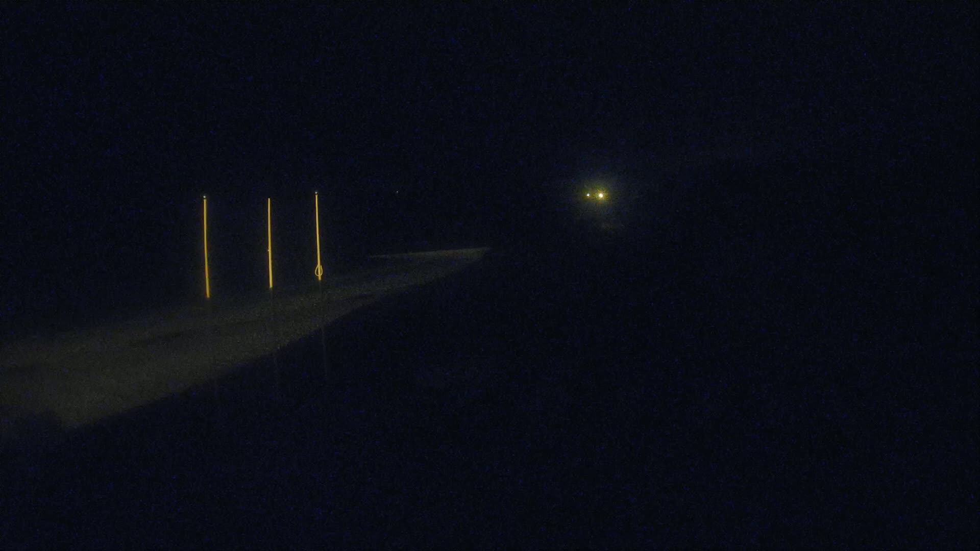 A dark, nighttime scene shows a road with three tall, thin, illuminated vertical posts alongside it, and a distant light source visible ahead.