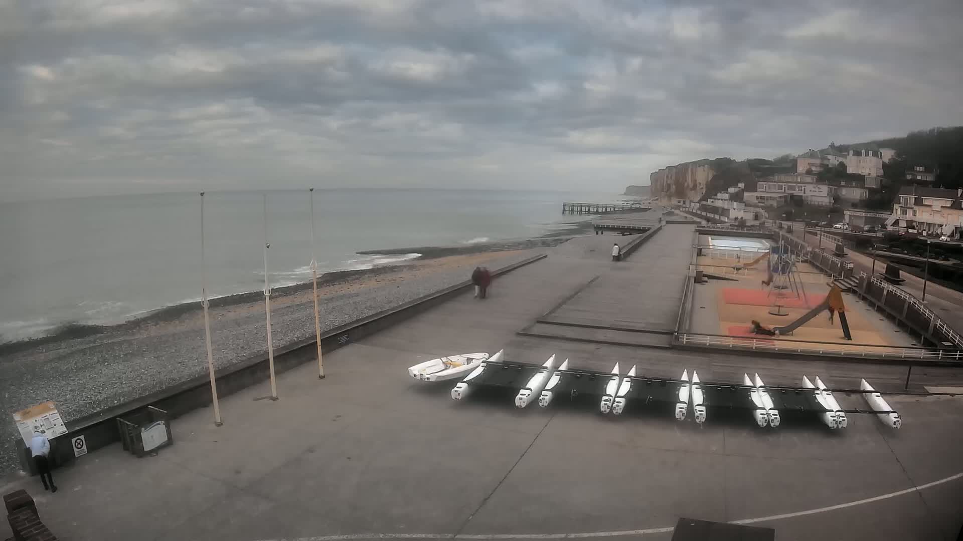 A gray, overcast day shows a seaside town with a pebble beach, a boardwalk, a playground, and several small sailboats lined up on the concrete.