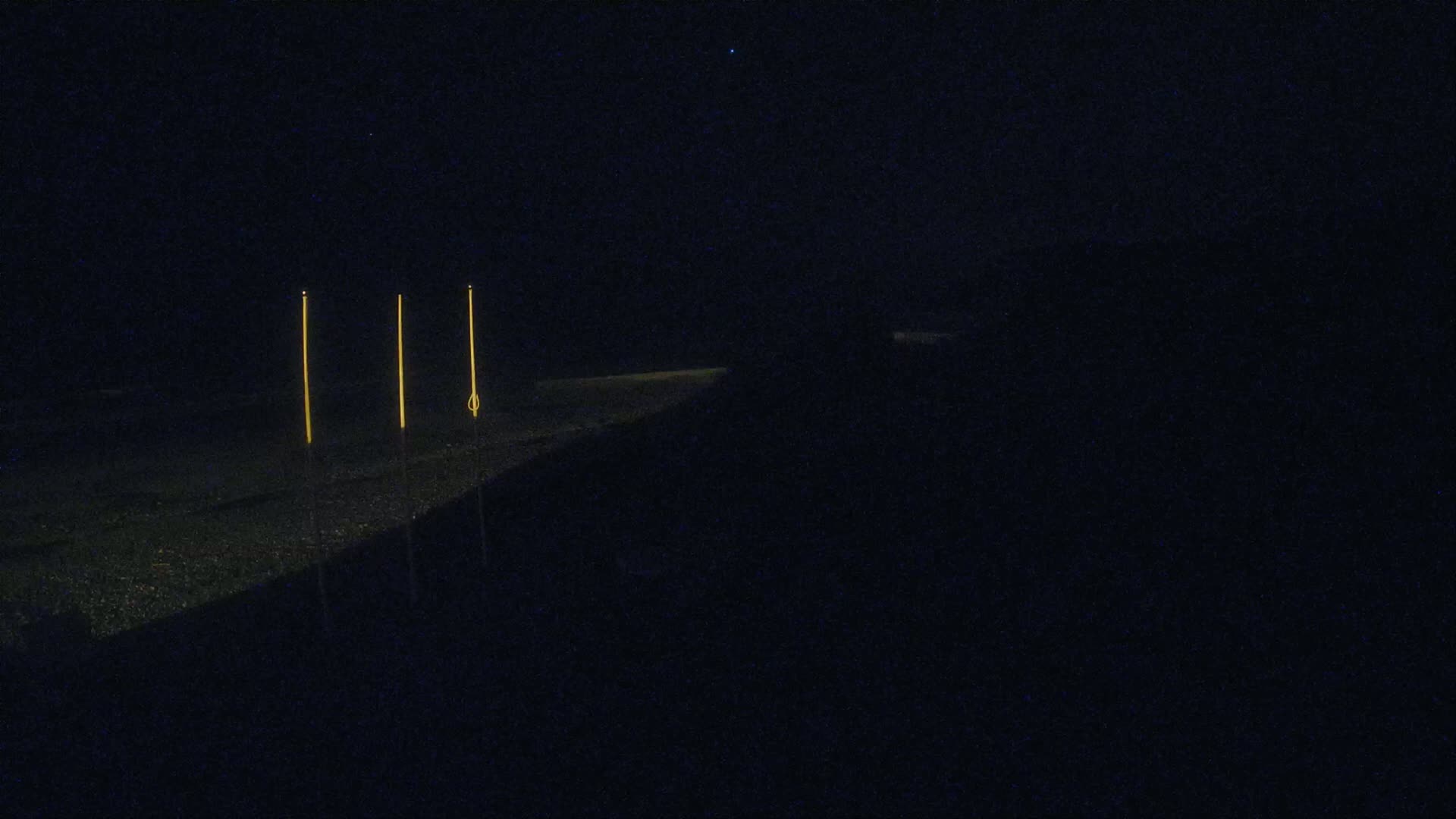 Three tall, thin, illuminated objects stand on a dark, possibly sandy surface at night under a clear, starlit sky.