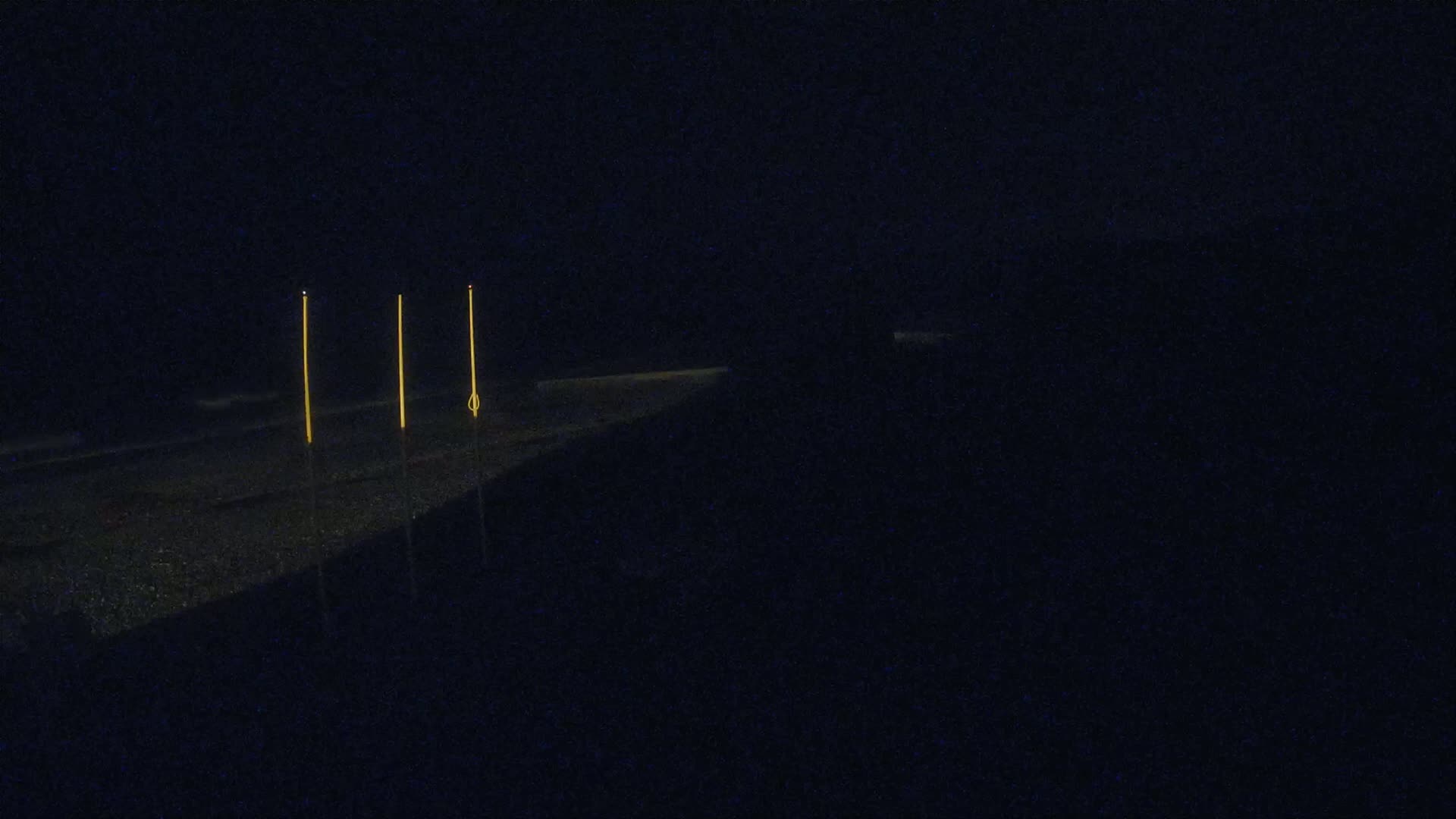 Three tall, thin, yellow lights stand on a dark, possibly asphalt, surface at night.