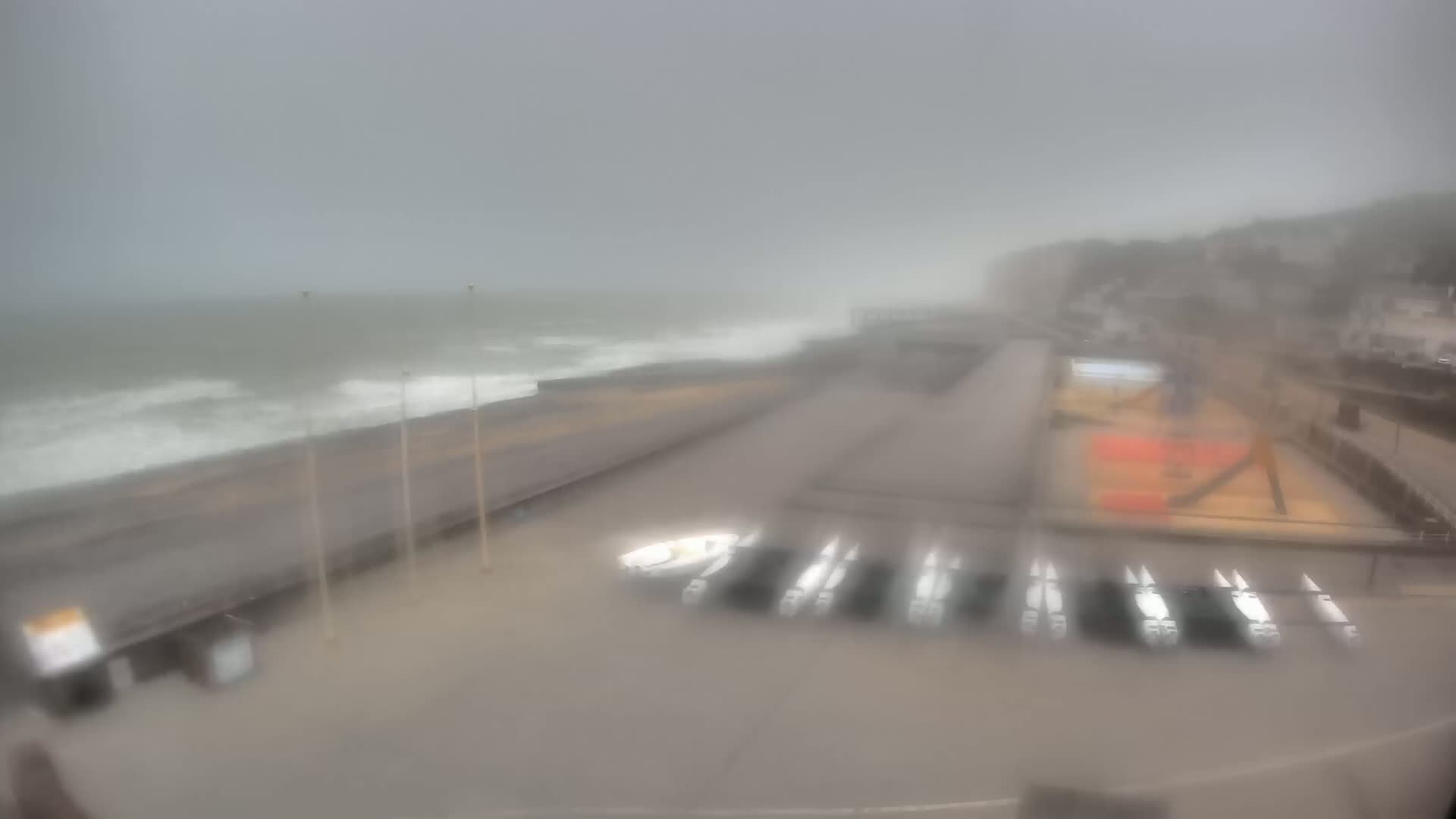 A rain-streaked view shows a coastal area with choppy waves crashing against a seawall, several small boats parked on the shore, and a hazy background of buildings and hills.