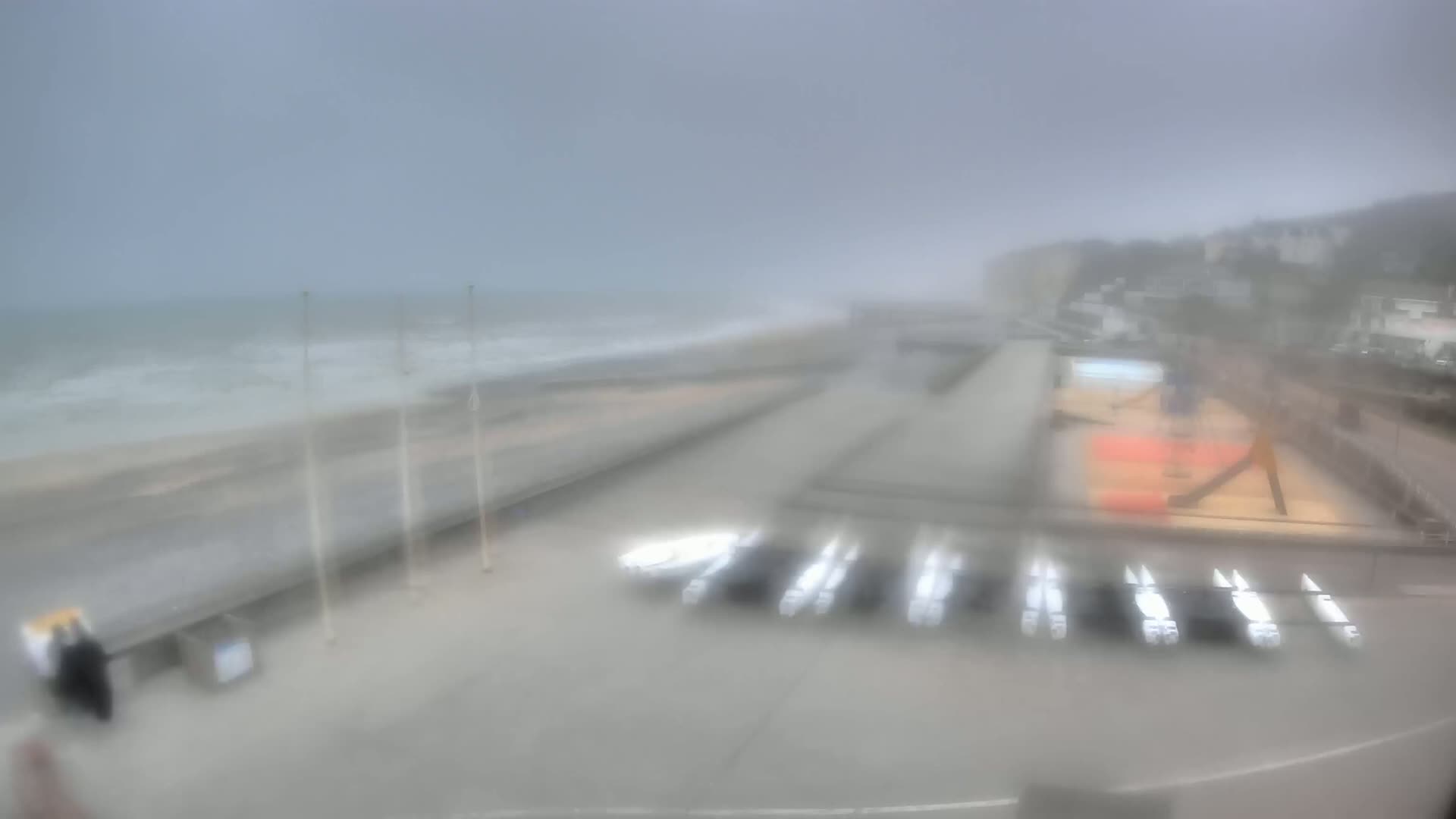 A blurry, overcast view of a beach with choppy waves, a boardwalk, and several boats stored on land.